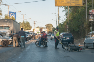 Gruppe von Menschen um ein verunglücktes Motorrad auf dem Seitenstreifen mit mehreren Fahrzeugen, darunter ein Lastwagen, im Hintergrund und Bäumen, Masten, Lichtern, Schildern und Himmel.