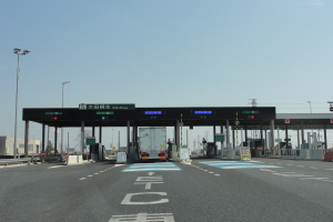 Eine Mautstelle mit einem geparkten Lastwagen auf einer Stra├če mit wei├čen und blauen Markierungen, Laternenmasten, Schildern und Geb├Ąuden im Hintergrund unter einem klaren blauen Himmel.