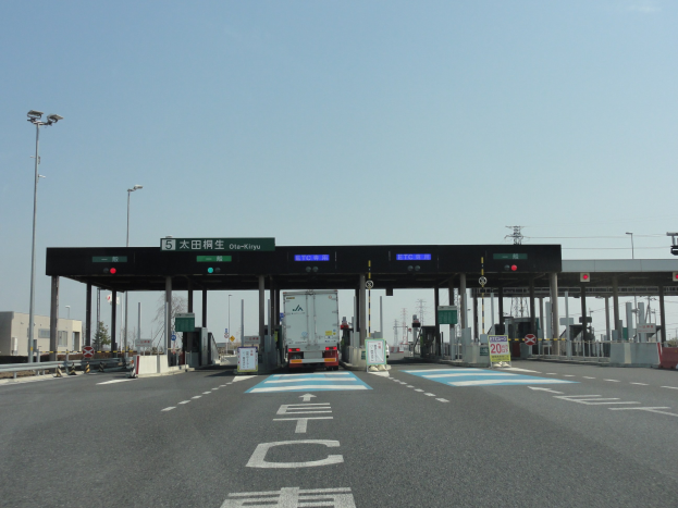 Eine Mautstelle mit einem geparkten Lastwagen auf einer Stra├če mit wei├čen und blauen Markierungen, Laternenmasten, Schildern und Geb├Ąuden im Hintergrund unter einem klaren blauen Himmel.