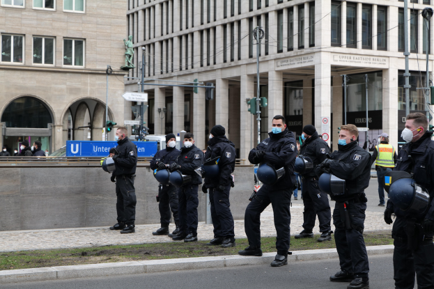 Eine Gruppe von Polizisten in schwarzen Uniformen und Masken steht vor einem Berliner Gebäude mit Glasfenstern und Säulen, einige halten Helme, mit Laternenmasten, Verkehrsampeln, Schildern mit Text, einer Statue und anderen Menschen im Hintergrund auf einem Rasen.