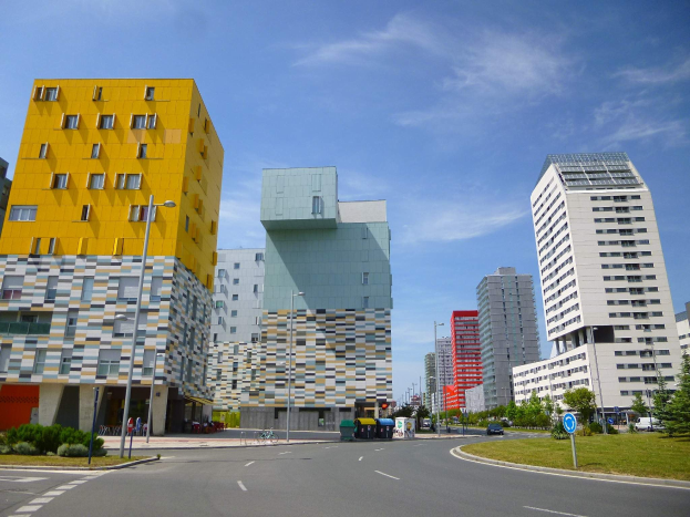 Eine Stadtstraße mit hohen Gebäuden im Hintergrund, einschließlich Strukturen der Universität Rotterdam, Straßenlaternen, Straßenlaternen, Kraftfahrzeugen, Informationsanzeigen, Büschen, Bäumen und einem bewölkten Himmel.