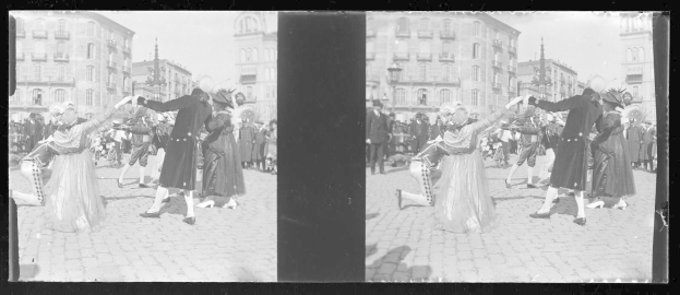 Schwarzes und weißes Foto einer Gruppe von Menschen in Kostümen, die auf der Straße während eines Karnevalsumzugs tanzen, umgeben von Gebäuden, Straßenlaternen und einer klaren Himmel.