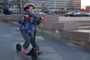 Ein junger Junge fährt mit einem Roller auf einem Gehweg, trägt einen Helm und Handschuhe, mit Stufen, Fahrzeugen, Menschen, Bäumen, Pfählen, Brettern, Gebäuden und einem klaren blauen Himmel im Hintergrund.