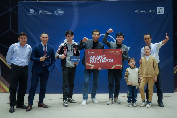 Gruppe von Menschen auf einer Bühne mit einer Tafel mit dem Text "Akang Kuchaydi - Zakovat Quiz", mit einem Banner, Lichtern und anderen Gegenständen im Hintergrund.