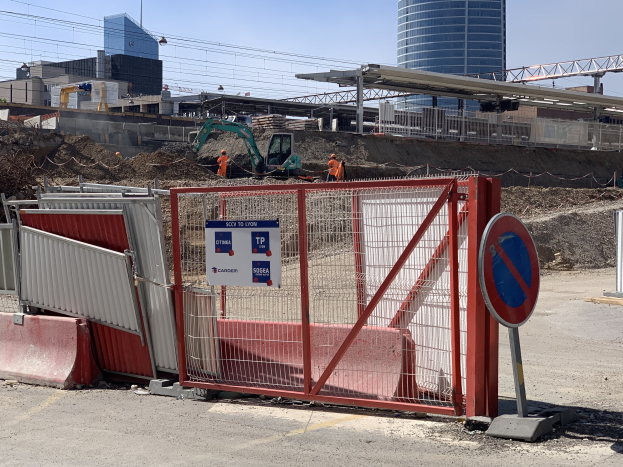 Baustelle mit rotem Tor, Metallzaun, Gebäuden, Kränen, Polen, Drähten und Menschen im Hintergrund bei klarem blauem Himmel.
