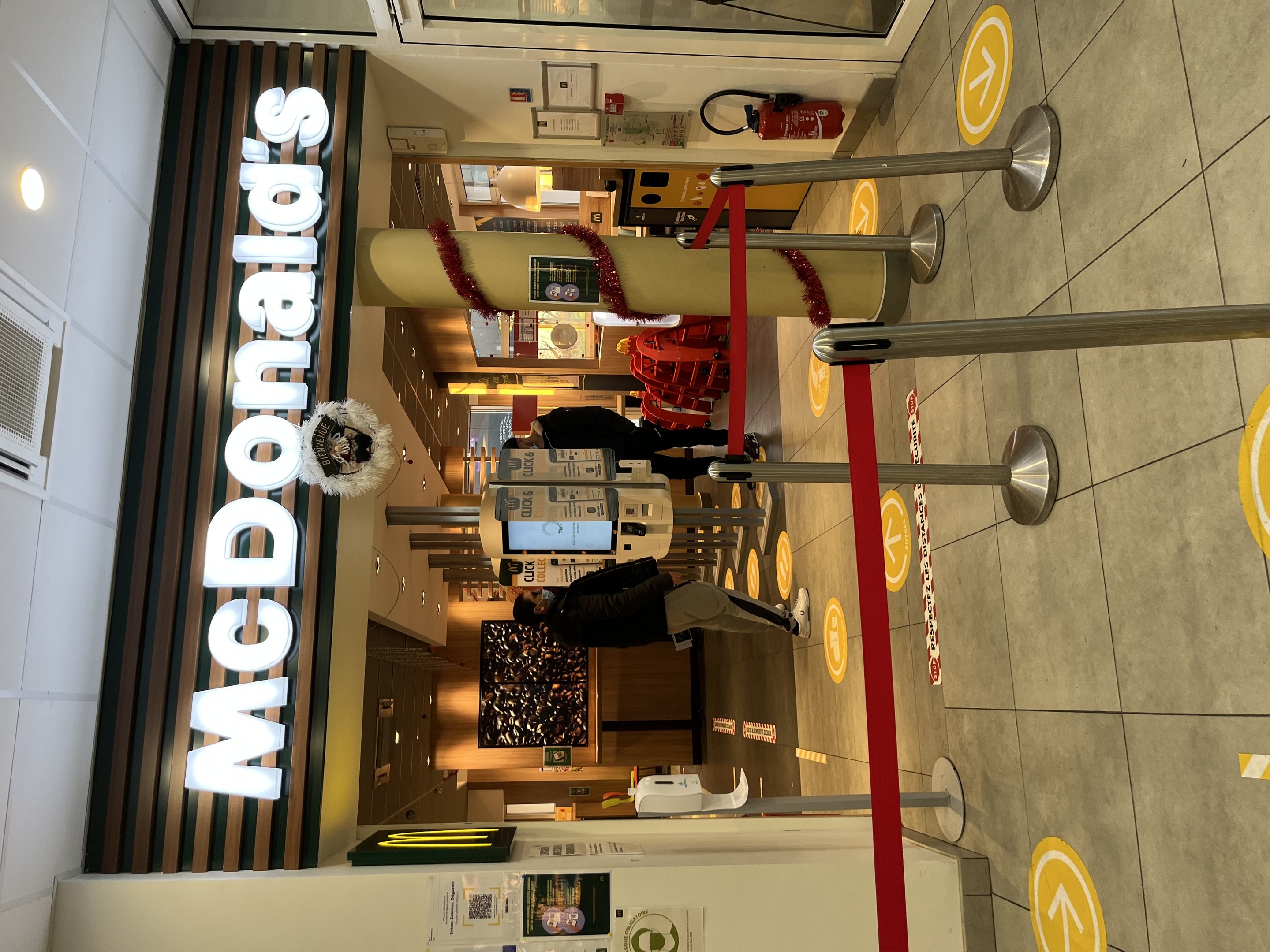 Innenseite eines McDonald's-Restaurants mit einem sichtbaren Schild, Metallstangen mit roten Bändern und einigen Menschen in der Nähe.