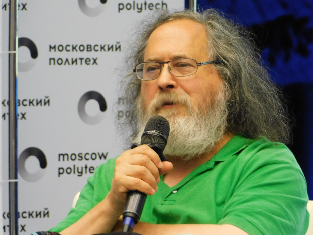 Ein Mann mit langen Haaren und Brille, der ein grünes T-Shirt trägt, sitzt auf einem Stuhl und hält ein Mikrofon, hinter ihm eine Tafel, die ihn als Psycholinguisten ausweist.