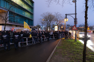 Eine große Gruppe von Menschen mit Schildern steht vor einem Gebäude, mit Barrikaden, Fahrrädern, Laternen, Schildern, Bäumen und Gras im Vordergrund und Himmel im Hintergrund, was auf eine Demonstration in Berlin hinweist.