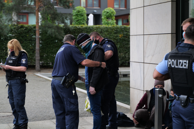 Deutsche Polizeibeamte stehen in einer Gruppe vor einem Gebäude, während eine Frau ein Buch und einen Stift hält und andere sitzen oder stehen in der Nähe von Grünflächen und städtischen Strukturen.
