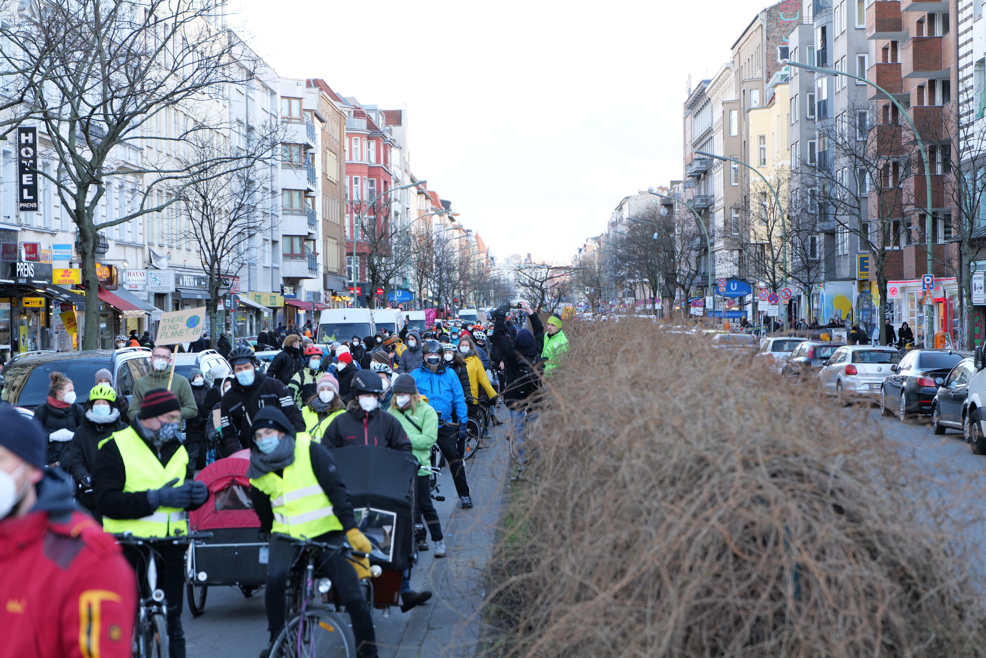 Eine große Gruppe von Menschen in Masken und Sicherheitswesten auf Fahrrädern eine von Bäumen gesäumte Straße mit Gebäuden, Laternen und Texttafeln entlang, neben Fahrzeugen und trockenem Gras, unter einem klaren blauen Himmel.