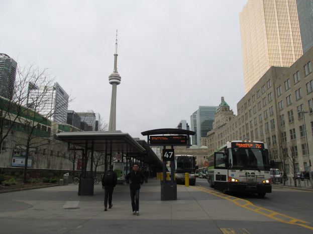 Eine belebte Stadtstraße mit einem Bus an einer Bushaltestelle vor einem hohen Gebäude, Fahrzeuge auf der Straße, Fußgänger auf dem Gehweg, ein überdachter Unterstand, Bäume, Gebäude mit Fenstern, ein Turm und der Himmel.