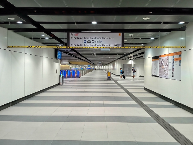 Ein langer Flughafenkorridor mit Menschen, Plakate an der rechten Wand, Deckenleuchten, eine Überwachungskamera und ein Gepäckabfertigungsschalter in der Mitte.