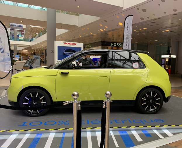 Ein gelbes wasserstoffbetriebenes Elektroauto vor einem Geb├Ąude geparkt, umgeben von Absperrpfosten mit Seilen, Bannern und Schildern, mit einer Person im Auto, einer Gruppe von Menschen in der N├Ąhe, einer S├Ąule, einem Topf mit einer Pflanze, Deckenleuchten und einer Decke mit Deckenventilatoren.