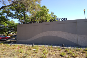 Außenansicht des Houstoner Holocaust-Museums mit eingravierter Textwand, Gedenksteine, Grünanlage, Straße mit Fahrzeugen, eine Straßenlaterne und eine bewölkte Himmel.