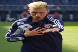 Ein junger Mann in einem blauen T-Shirt steht auf einem Fußballfeld mit ausgestreckten Armen und entschlossener Miene, mit einem unscharfen Hintergrund.