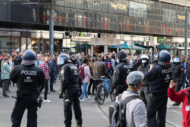 Eine Gruppe von Polizeibeamten, die vor einer Menschenmenge auf der Straße stehen, wobei einige Personen Helme tragen und Taschen oder Fahrräder haben, und Gebäude mit Glaswänden und Schildern im Hintergrund.