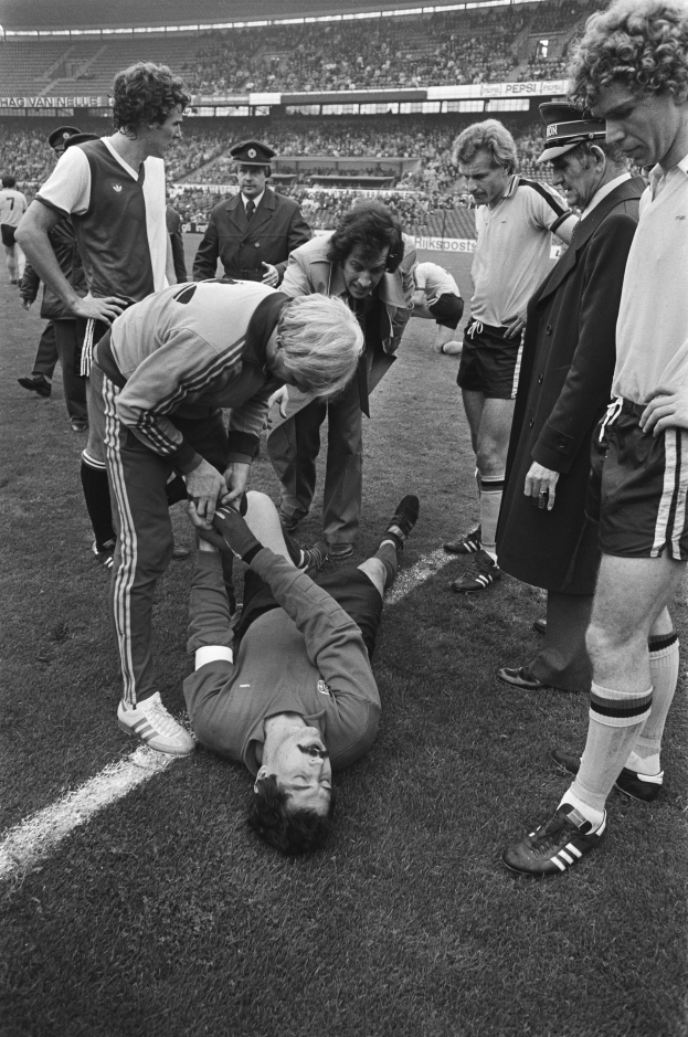 Schwarzes und weißes Foto eines Füßballspielers, der auf dem Boden liegt und von einer Gruppe von Männern umgeben ist, mit einer Stadionmenge im Hintergrund; der Spieler sieht verletzlich aus.