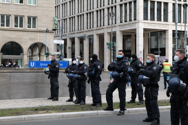 Eine Gruppe von Polizisten in schwarzen Uniformen und Masken steht vor einem Glasfenstergebäude mit Säulen in Berlin, Deutschland, einige halten Helme, mit Laternenmasten, Verkehrsampeln, Schildern mit Text, einer Statue, anderen Menschen und grasbewachsenem Boden im Hintergrund.