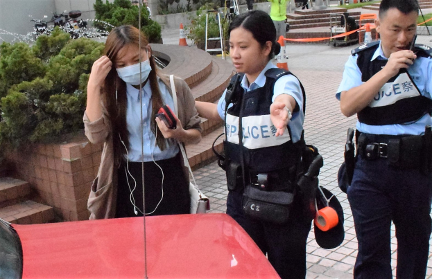 Zwei Polizisten sprechen mit einer Frau vor einem roten Auto, mit Grünpflanzen und einem Gebäude im Hintergrund.
