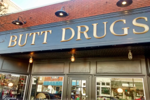 Außensicht eines Gebäudes mit einer beleuchteten Schilder 'butt drugs' und gläsernen Wänden, die umliegende Fahrzeuge, Gebäude, Bäume und Himmel zeigen.