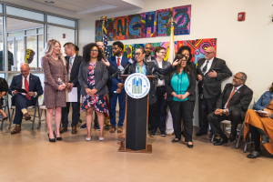 Gruppe von Menschen in einem Raum mit einem Podium, Mikrofon und einer Tafel mit der Aufschrift "Sancho-Stadtrat tritt dem Stadtrat bei", umgeben von Fahnen, Stühlen und Deckenleuchten.