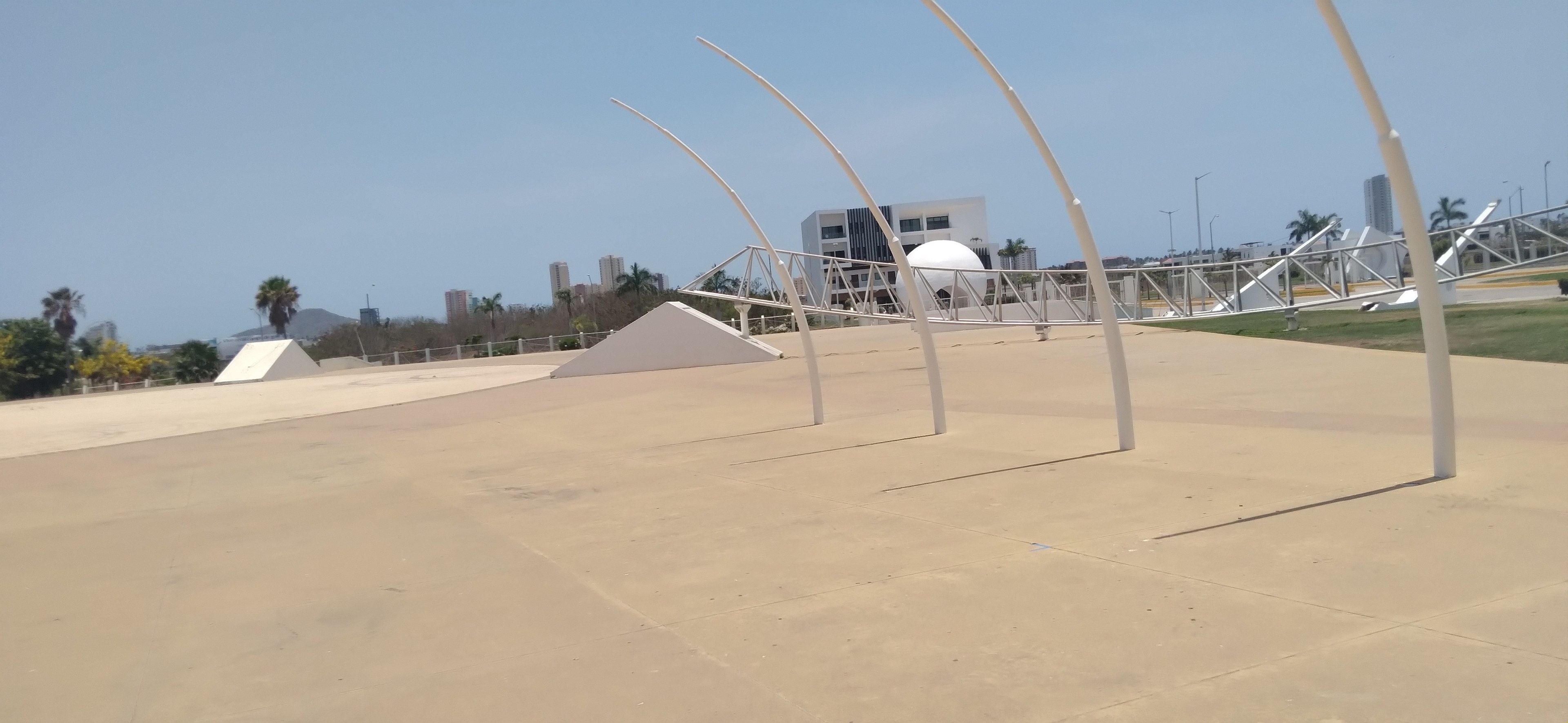 Ein Skaterpark mit einer zentralen großen Metallstruktur, umgeben von Pfosten, Gras, einem Zaun, Bäumen, Gebäuden, Straßenlaternen und einem bewölkten Himmel.