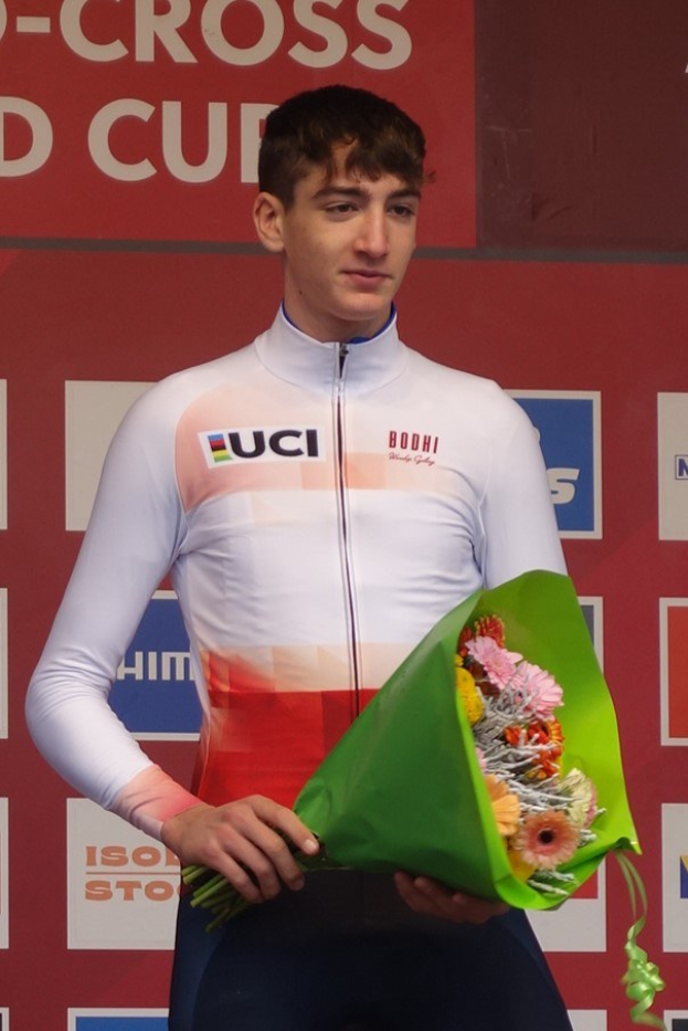 Ein Mann steht triumphierend auf einem Podium, hält einen Blumenstrauß in der Hand und hat ein Schild im Hintergrund, strahlt Freude und Selbstbewusstsein aus.