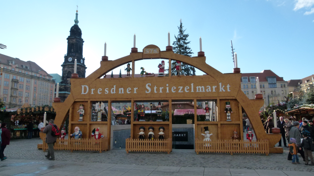 Ein lebendiger Weihnachtsmarkt in Dresden, Deutschland, mit Menschen, die herumlaufen, einem Bogen mit Text, Tannenbäumen, Gebäuden mit Fenstern und einem bewölktem Himmel.
