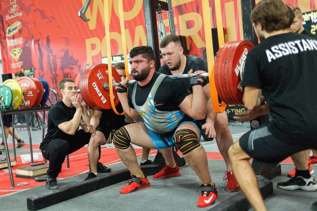 Eine Gruppe von Männern führt Kniebeugen mit Hanteln in einem Fitnessstudio durch, das mit Equipment und Bannern ausgestattet ist, auf denen "Powerlifting-Weltmeisterschaften 2019" steht.