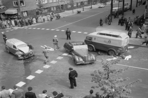 Eine Schwarz-Weiß-Szene eines Autounfalls an einer Straßenecke mit mehreren Fahrzeugen, darunter ein Lieferwagen, und einer Gruppe von Menschen, die herumstehen, mit Gebäuden, Bäumen, Masten und Texttafeln im Hintergrund.