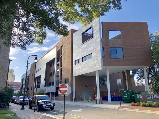 Großes mehrstöckiges Stadtzentrumsgebäude mit zahlreichen Fenstern, Straßeninfrastruktur einschließlich Pfosten, Lampen und Verkehrsmanagementmerkmale, umgeben von Grünflächen und Fahrzeugen an einem bewölkten Tag.