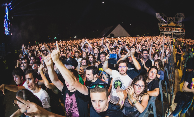 Eine große Menschenmenge auf einem Musikfestival mit erhobenen Händen, sitzend und stehend, umgeben von einem Metallzaun, Gebäuden und Lichtern im Hintergrund unter einem dunklen Himmel, mit einem Mann und einer Frau im Vordergrund.