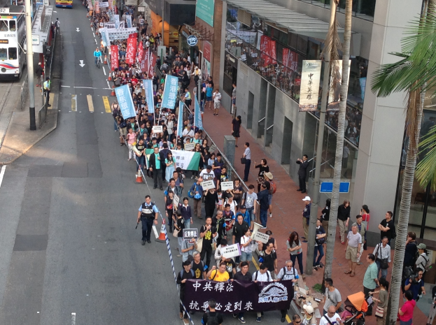 Eine große Gruppe von Menschen marschiert durch eine Straße in Hong Kong und hält Protestschilder hoch, wobei Bäume, Glasfassaden, Gebäude und Fahrzeuge zu sehen sind.