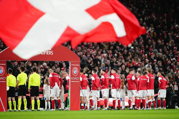 Eine Gruppe von Menschen auf einem Fußballfeld mit einer roten und weißen Flagge im Vordergrund, einem Bogen mit 'Bayern Munich vs Bayern Munich Betting Tips & Preview' im Hintergrund und einer großen Menge im Stadion.