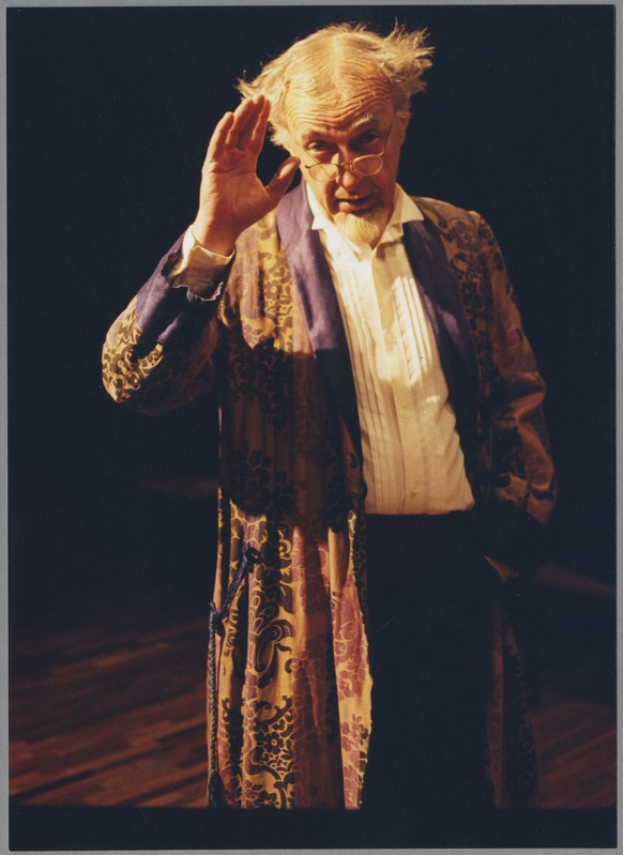 Ein Mann in einem Gewand und Brille steht auf einer Bühne mit dunklem Hintergrund, die Hände zusammenlegend und ernst in die Menge schauend.