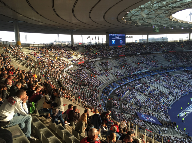Eine große Menschenmenge in einem Stadion schaut einem Fußballspiel zu, mit einer Bühne, Fahnen und einem Bildschirm im Hintergrund, wahrscheinlich im Allianz Arena in München, Deutschland.