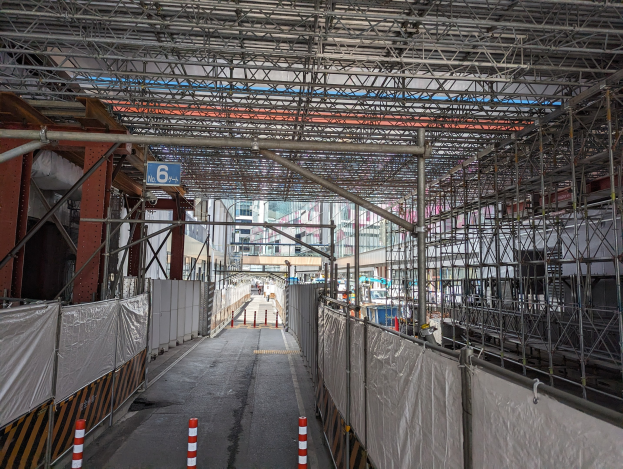 Baustelle mit ausgedehnten Gerüsten, einem Zaun, einem Schild, Metallrahmen, benachbarten Gebäuden und einem bewölkten Himmel.