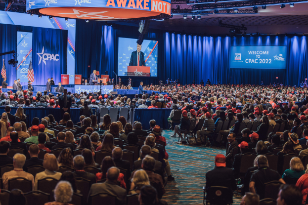 Eine große Menschenmenge sitzt auf Stühlen, die einer Bühne zugewandt sind, mit ein paar Menschen, die auf dem Podium und auf der linken Seite stehen, blaue Vorhänge und ein Bildschirm im Hintergrund und ein orangefarbenes Schild oben, das die CPAC-Veranstaltung am 25. Oktober anzeigt.