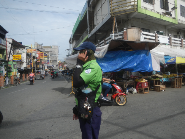 Ein Polizeibeamter in grüner und blauer Uniform und blauer Mütze steht auf der Seite einer Straße und spricht auf einem Handy, mit vorbeifahrenden Fahrzeugen, Ständen mit Gegenständen rechts von ihm und einem bewölkten Himmel im Hintergrund.