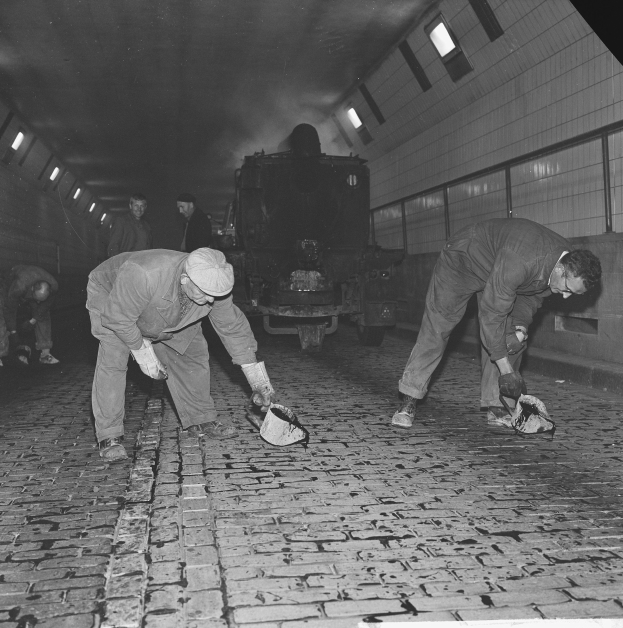 Schwarzes und weißes Bild von Männern, die auf einem Ziegelboden in einem Tunnel arbeiten, mit einem Zug im Hintergrund, der von Deckenlampen beleuchtet wird.