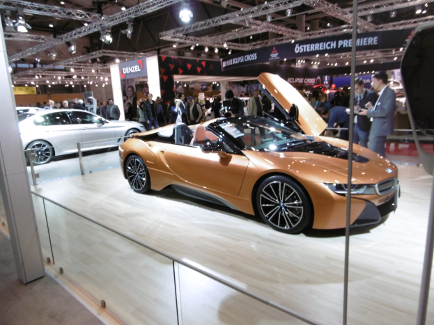 Ein schwarzer BMW i8 Roadster mit gl├Ąnzendem Finish und Chromakzenten auf der Frankfurt Motor Show umgeben von formell gekleideten G├Ąsten in einer Atmosph├Ąre der Aufregung.