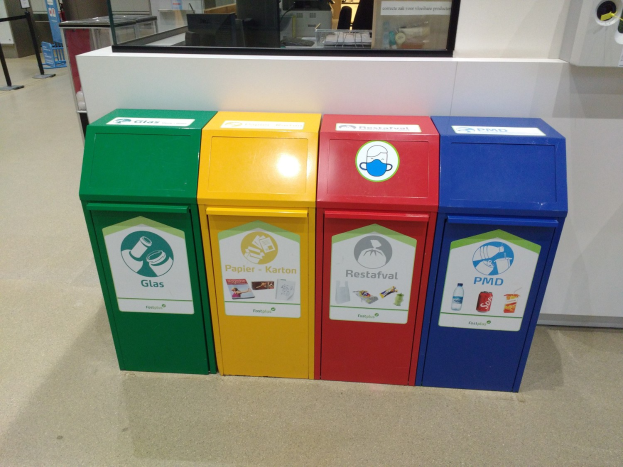 Vier bunte Mülltonnen in einer Reihe, mit einer Arbeitsplatte, einem Monitor und anderen Gegenständen im Hintergrund und einer Person links daneben.