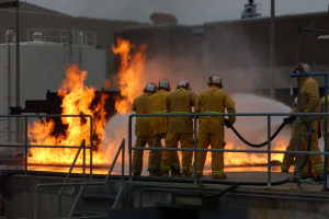 Eine Gruppe von Feuerwehrleuten in Uniform steht zusammen in einem Raum mit einer Tür auf der rechten Seite und einer Wand auf der linken Seite, mit Rohren und anderen Gegenständen im Hintergrund.