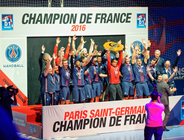 Männergruppe auf einer Bühne mit einem Pokal, einer Person, die filmt, und einem 'Champion de France 2016'-Schild im Hintergrund.