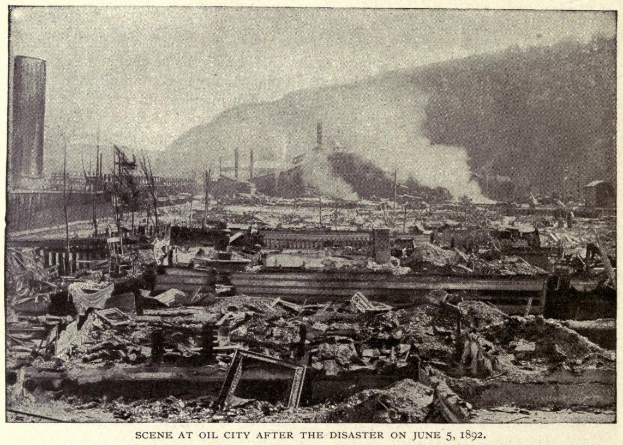 Schwarzes und weißes Foto einer Stadt nach einer Katastrophe am 5. Juni 1892, das Gebäude, Pfosten, Rauch, einen Hügel im Hintergrund und Text am unteren Rand zeigt.