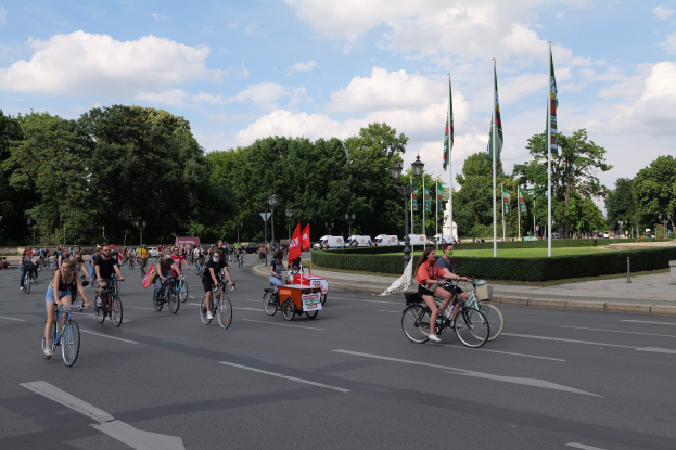 Eine Gruppe von Menschen, die auf Fahrrädern eine von Bäumen gesäumte Straße entlangfahren, während des Paris Bike Ride 2016. Im Hintergrund sind Laternenmasten, Fahnenmasten, eine bewölkte Himmel und Fahrzeuge zu sehen.