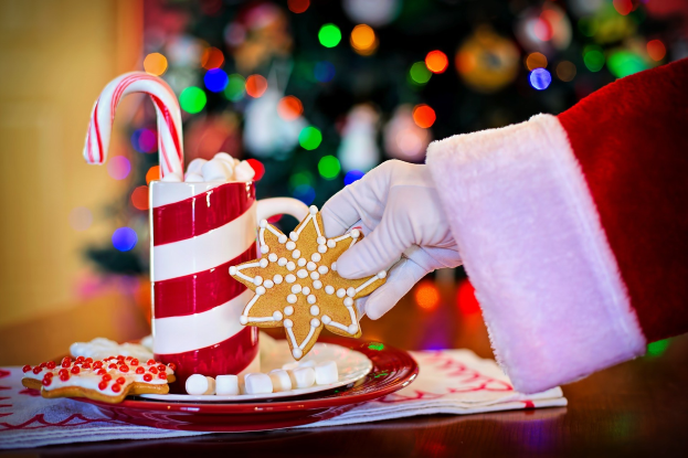 Eine Person mit Santa Claus Mütze, die einen Becher heißen Kakao und einen Zuckerstange hält und ein Teller mit Keksen und ein Tuch auf dem Tisch vor ihnen liegen hat, vor einem Weihnachtsbaum und einer Wand.