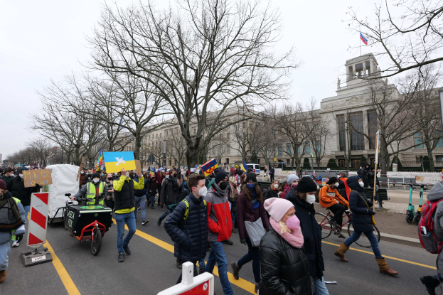 Eine große Protestmarsch mit Menschen, die eine Straße in Washington, D.C. entlanggehen, einige halten Schilder und fahren Fahrräder, unter einem klaren blauen Himmel mit Bäumen und Schildern im Hintergrund.