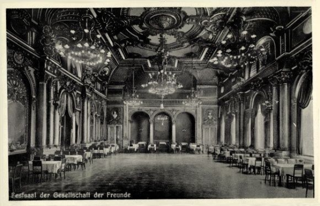 Schwarz-weißes Foto eines großen Raums mit Tischen, Stühlen, Säulen, Bögen und Kronleuchtern, beschriftet mit "Festival der Gesellschaft der Freunde, Berlin" unten.