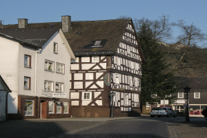 Half-timbered house in a small town surrounded by buildings, vehicles, pedestrians, trees, and street infrastructure under a clear blue sky.